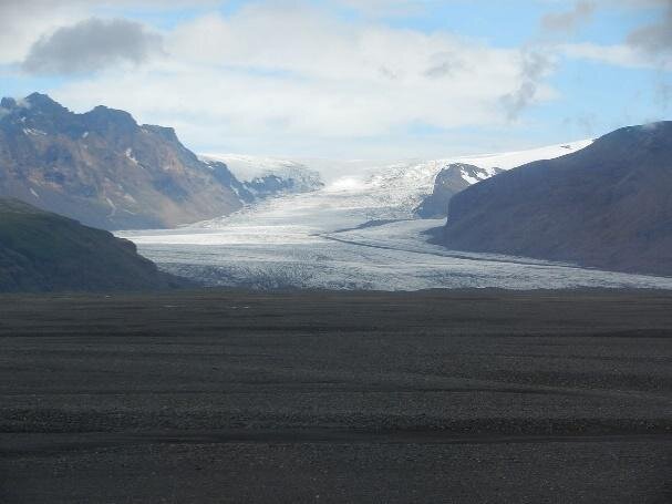 Na pierwszym planie fotografii rozległy, niepokryty roślinnością teren o ciemnoszarej barwie. W tle schodzący ze stoku wzniesienia szeroki lodowiec. Po bokach inne wzniesienia.