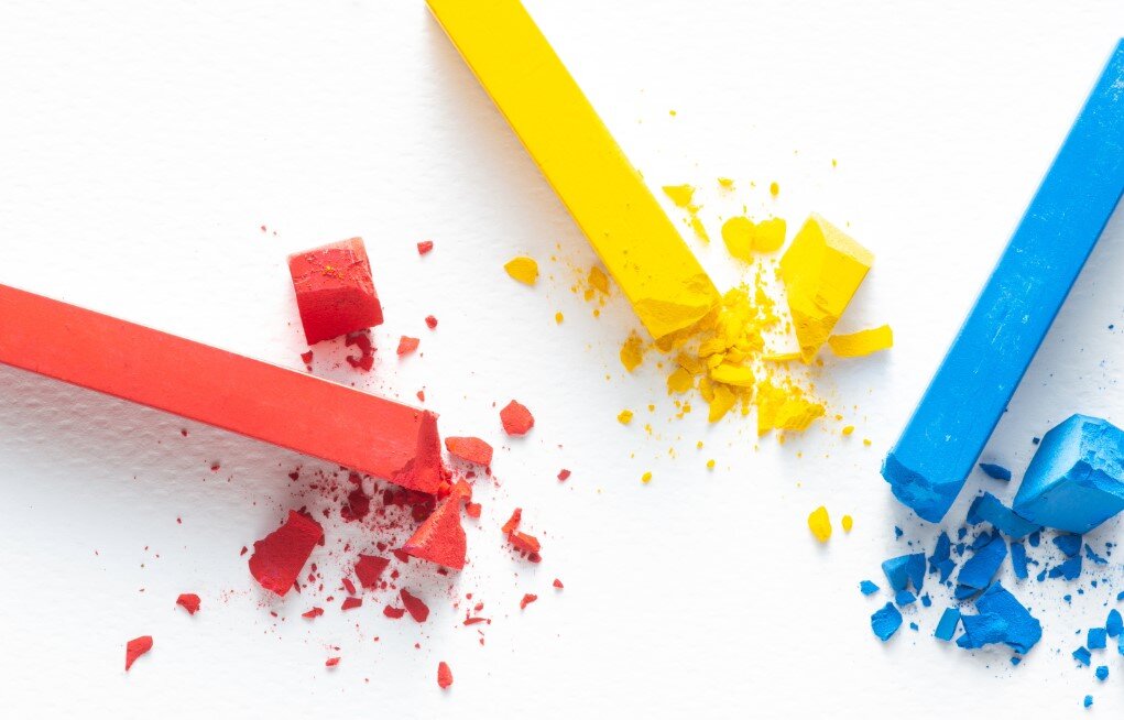Ilustracja przedstawia trzy kolorowe kredy ułożone obok siebie, od lewej czerwona, żółta i niebieska. Obok każdej kredy leżą pokruszone kawałki. Tło jest białe.