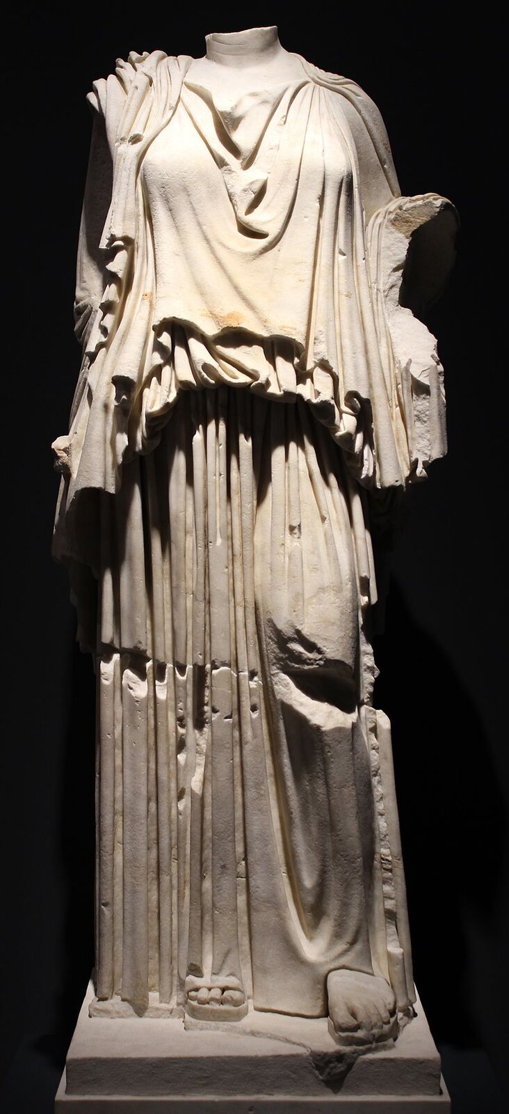 Zdjęcie przedstawia posąg w białym kolorze na czarnym tle. Jest to żeńska postać bez głowy, ubrana w luźną szatę, związaną w pasie. Ma bose stopy.