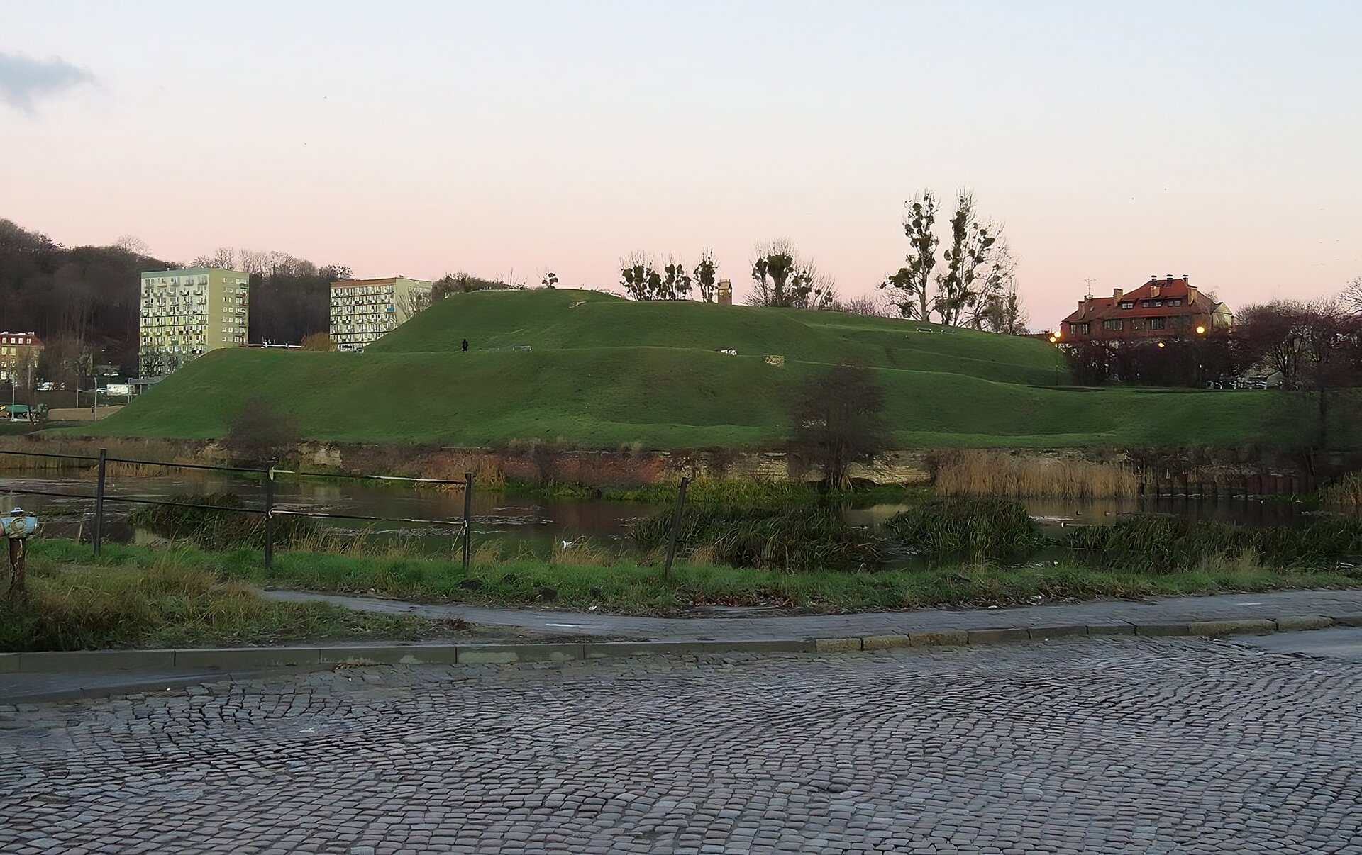 Zdjęcie przedstawia widok współczesny jednego z bastionów fortyfikacji nowożytnego Gdańska - bastion św. Gertrudy. Nasyp ziemny, zwężany do szczytu w dwóch rzędach, od strony zewnętrznej wzmocniony poprzez ceglane umocnienia. Na nasypach rampy wjazdowe usypane na potrzeby artylerii obronnej. W tle wzgórza  współczesne zabudowania bloków mieszkalnych, na pierwszym planie brukowana droga.