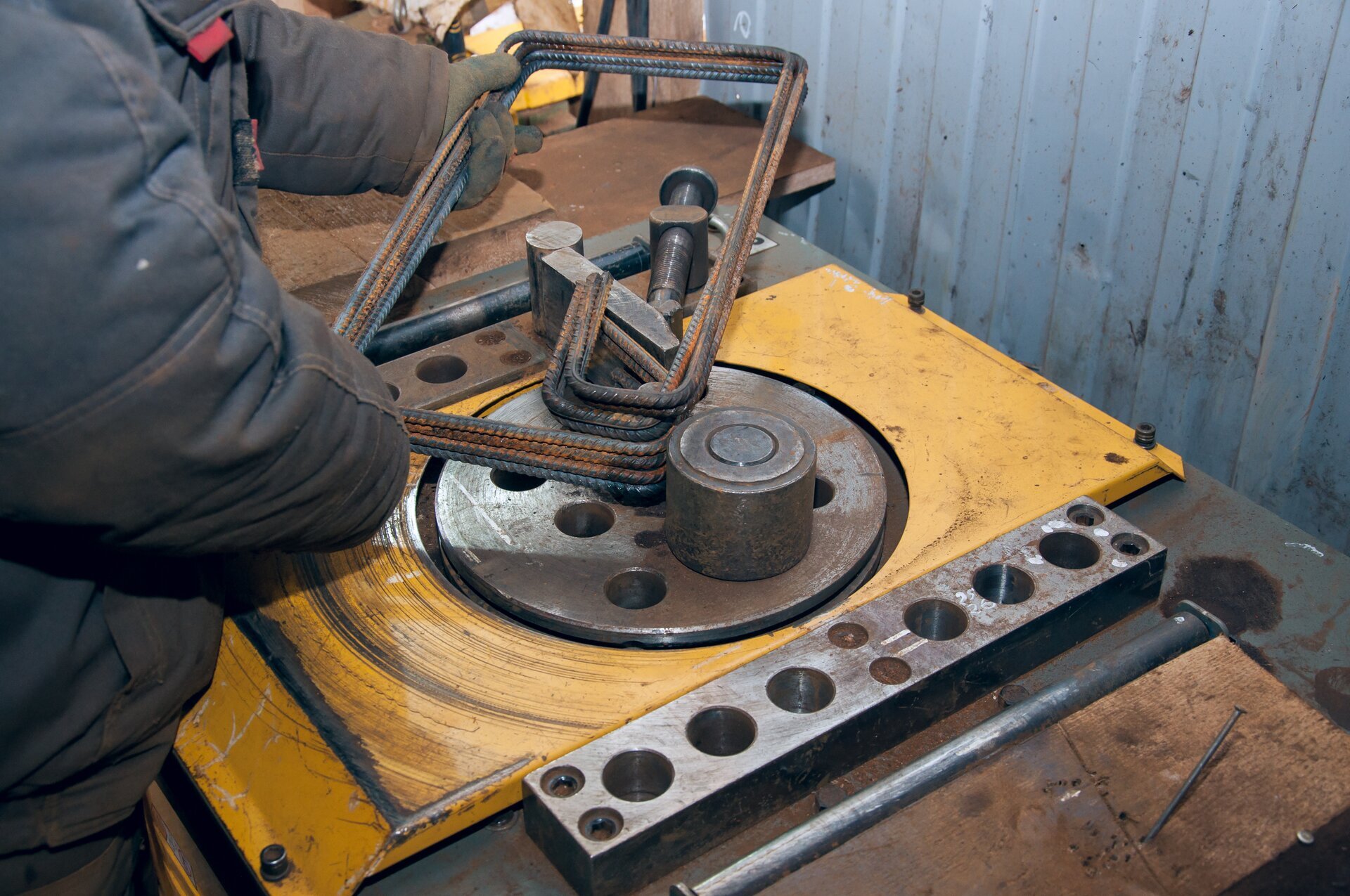 Zdjęcie przedstawia giętarkę mechaniczną o żółtej budowie. Robotnik trzyma kilka prętów, które wkłada w mechanizm maszyny i odgina.