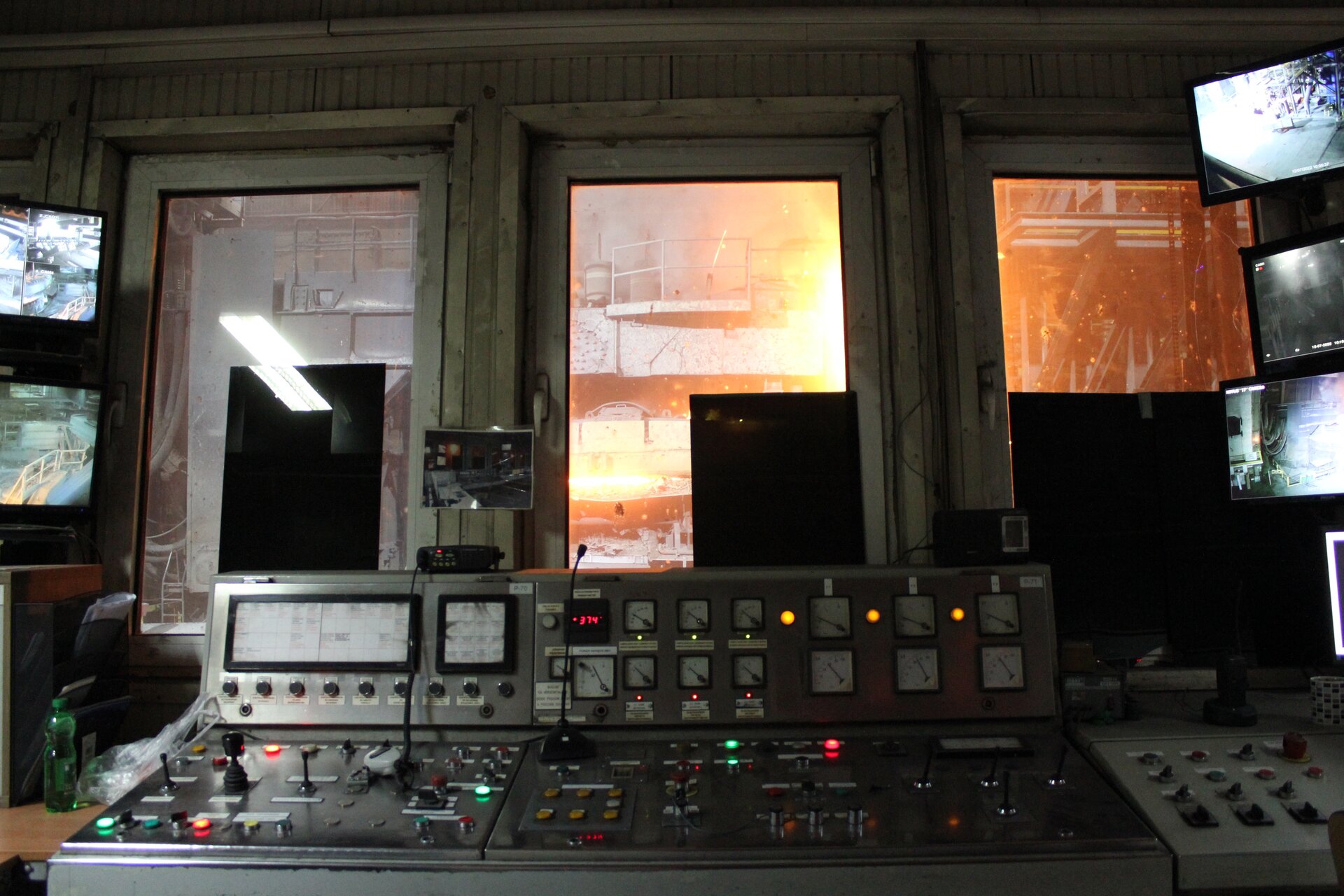 Zdjęcie przedstawia pulpit sterowniczy w kabinie operatorskiej elektrycznego pieca łukowego. Na pierwszym planie widoczny jest pulpit sterowniczy. Znajduje się na nim duża ilość przycisków, dźwigni oraz tarcz manometrów. Dookoła widoczne są monitory. Część z nich jest wyłączona, a na części widoczny jest obraz z kamer. Na drugim planie widoczne są trzy okna. Przez okna widać metalową konstrukcję, balkony, schody oraz iskrę.