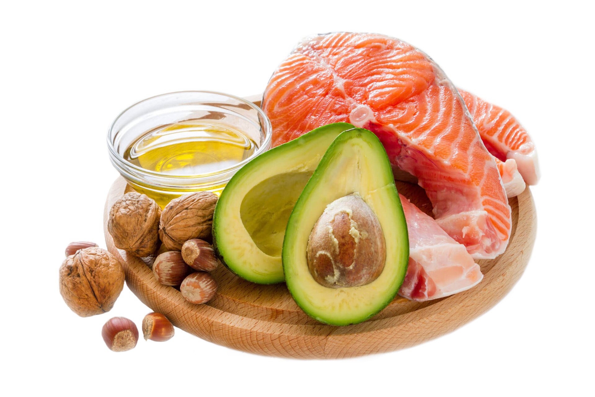 Zdjęcie przedstawia przykładowe produkty dostarczające tłuszczy. Na drewnianej okrągłej desce widoczne są: dwa dzwonka łososia, przecięte na pół awokado, trzy orzechy włoskie i pięć orzechów laskowych, szklana miska z żółtym olejem.
