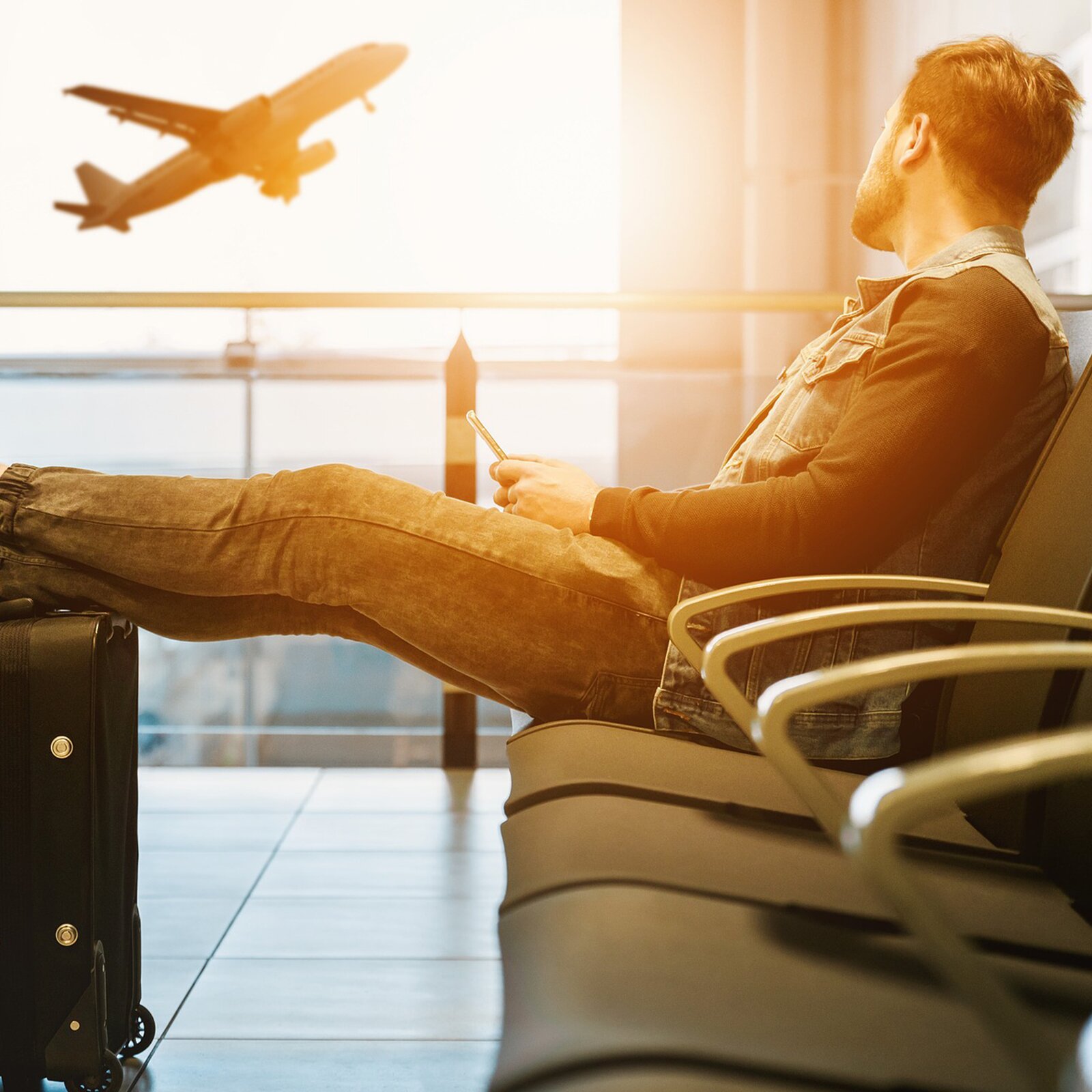 Zdjęcie przedstawia mężczyznę siedzącego w hali lotniska z nogami opartymi o walizkę. W tle w oknie widoczny jest startujący samolot.
