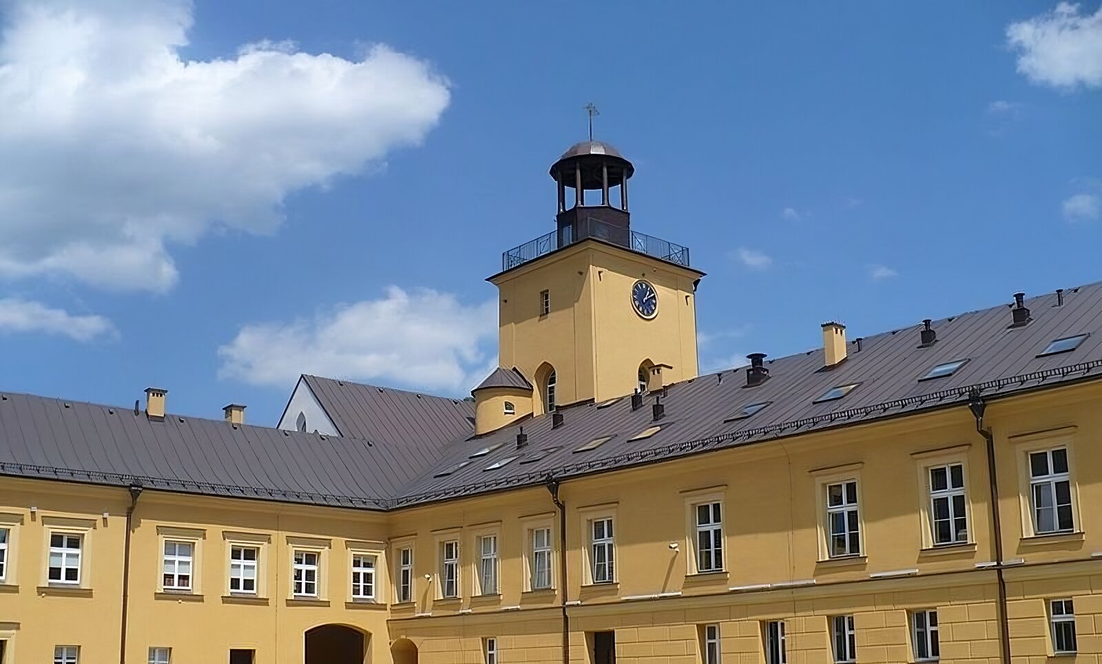 Zdjęcie przedstawia widok współczesny na fragment zabudowy pałacu w Koszęcinie. W centralnej części fotografii, w lewym skrzydle budynku znajduje się łączenie dwóch skrzydeł gmachu z portalem bramnym u dołu. Powyżej widoczna wieża zegarowa o kształcie sześcianu. Na szczycie wieży znajduje się balustrada i mniejsza kopuła wsparta na czterech filarach. W centrum fotografii, na lewo od wieży zegarowej, widoczny fragment walcowatej wieżyczki z okienkiem, dobudowanej do wieży głównej. Skrzydło gmachu po prawej stronie fotografii posiada na dachu otwory wentylacyjne i okna połaciowe. Na całej długości fotografii ukazano linie okien na piętrze i na parterze. Fasada obiektu - ściany, gzymsy, fryzy i obramowania okien - wykonane w kolorze żółtym. 