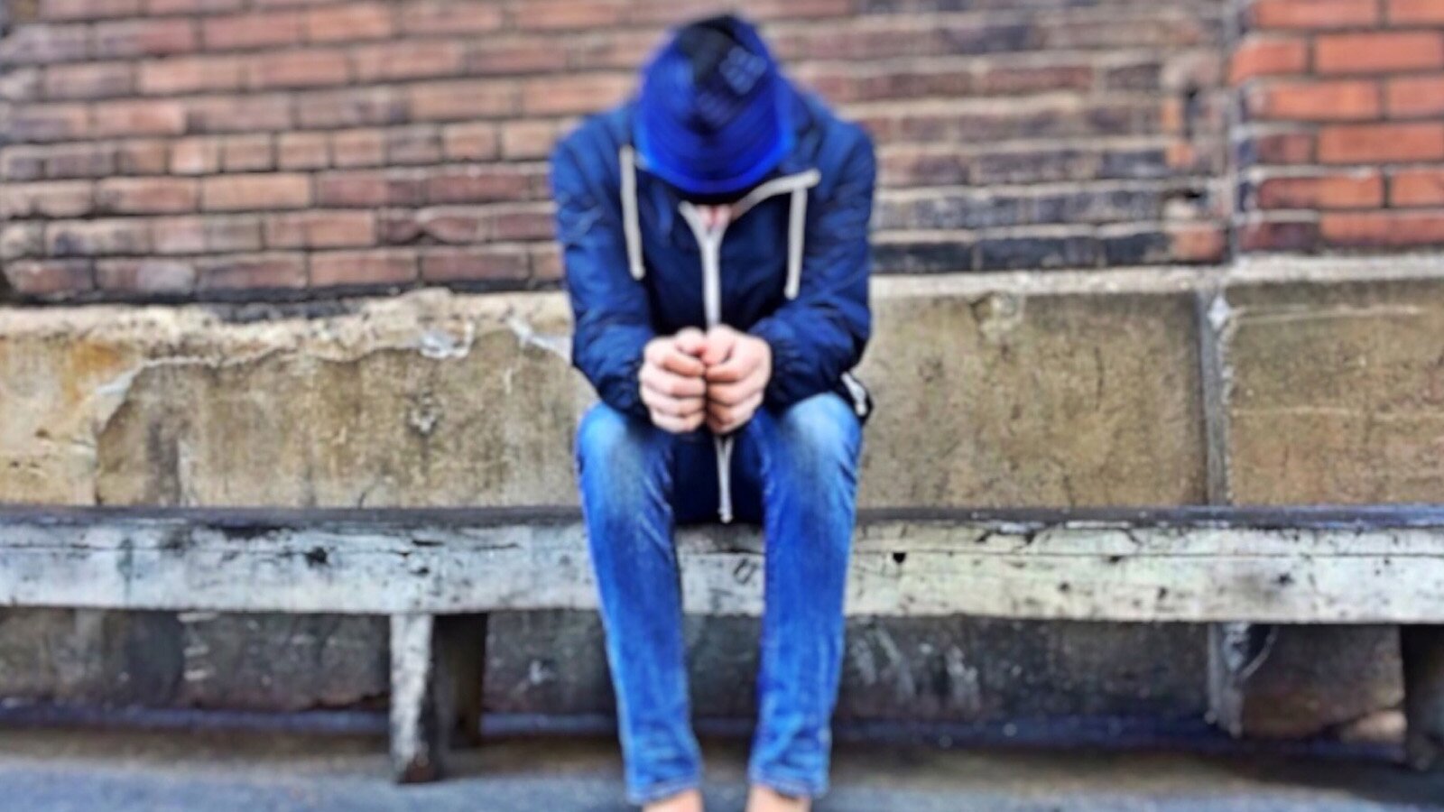 Zdjęcie przedstawia siedzącego na murku człowieka. Głowę ma pochyloną i dłonie zaciśnięte w pięści.