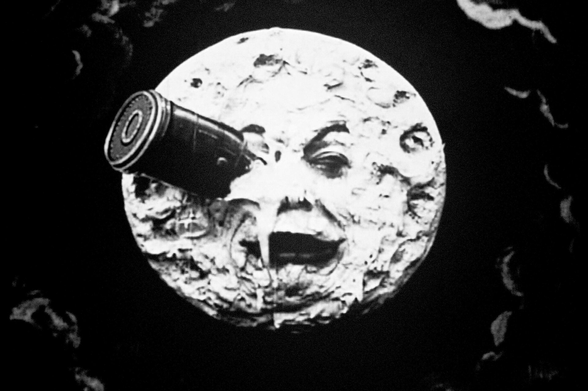 Ilustracja przedstawia kadr z filmu „Podróż na Księżyc” autorstwa Georgesa Mélièsa. Na czarno-białym zdjęciu znajduje się okrągły, pełen kraterów księżyc o ludzkiej twarz i dużych, ciemnych ustach, w którego prawym oku tkwi wbita metalowa rakieta. Jasna tafla księżyca unosi się na tle czarnego nieba z zaznaczonymi gdzieniegdzie szarymi zarysami chmur.