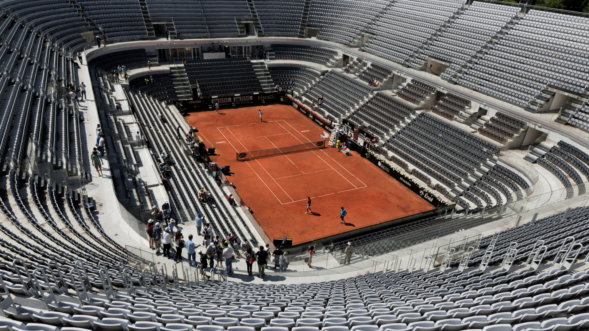 Zdjęcie przedstawia stadion tenisowy. W dole widoczny jest kort, a wokoło puste trybuny.