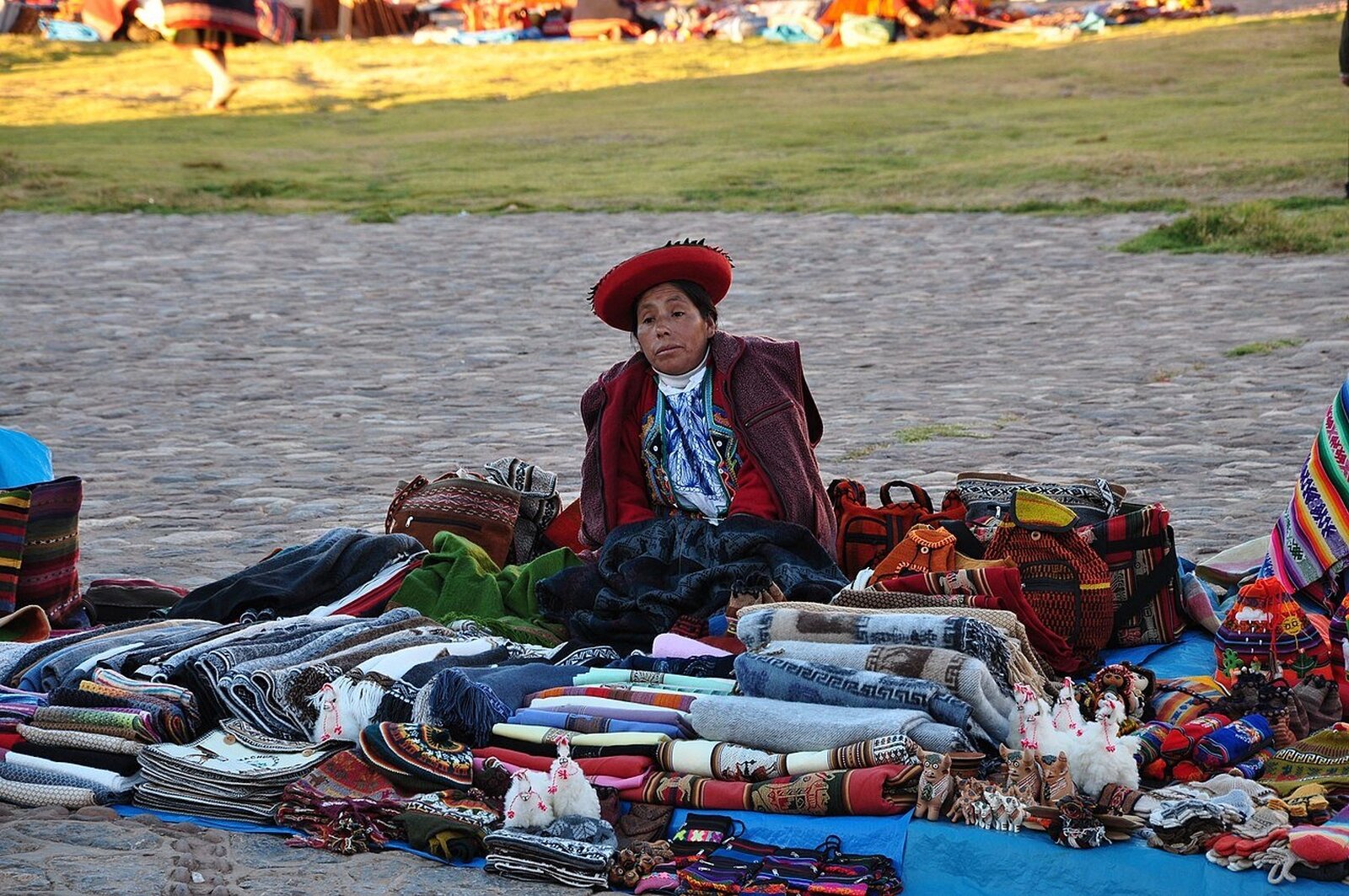 Zdjęcie przedstawia Indiankę siedzącą na ziemi wyłożonej kamieniami, mająca przed sobą ręcznie wykonane koce, torby, maskotki i podobne wyroby. Są bogato zdobione, w różnych kolorach i wzorach.