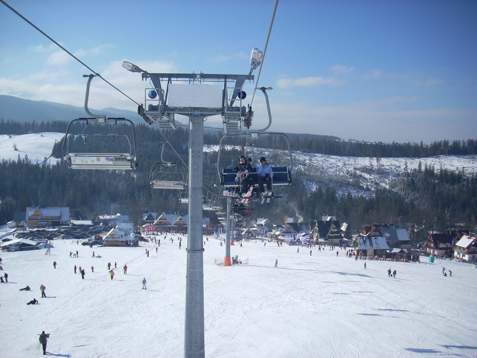 Fotografia przedstawia wyciąg krzesełkowy zimą. Na nim po prawej stronie osoby wjeżdżające na szczyt z nartami. Poniżej pokryta śniegiem powierzchnia, na której są osoby zjeżdżające na nartach. Wokół drewniane chatki.