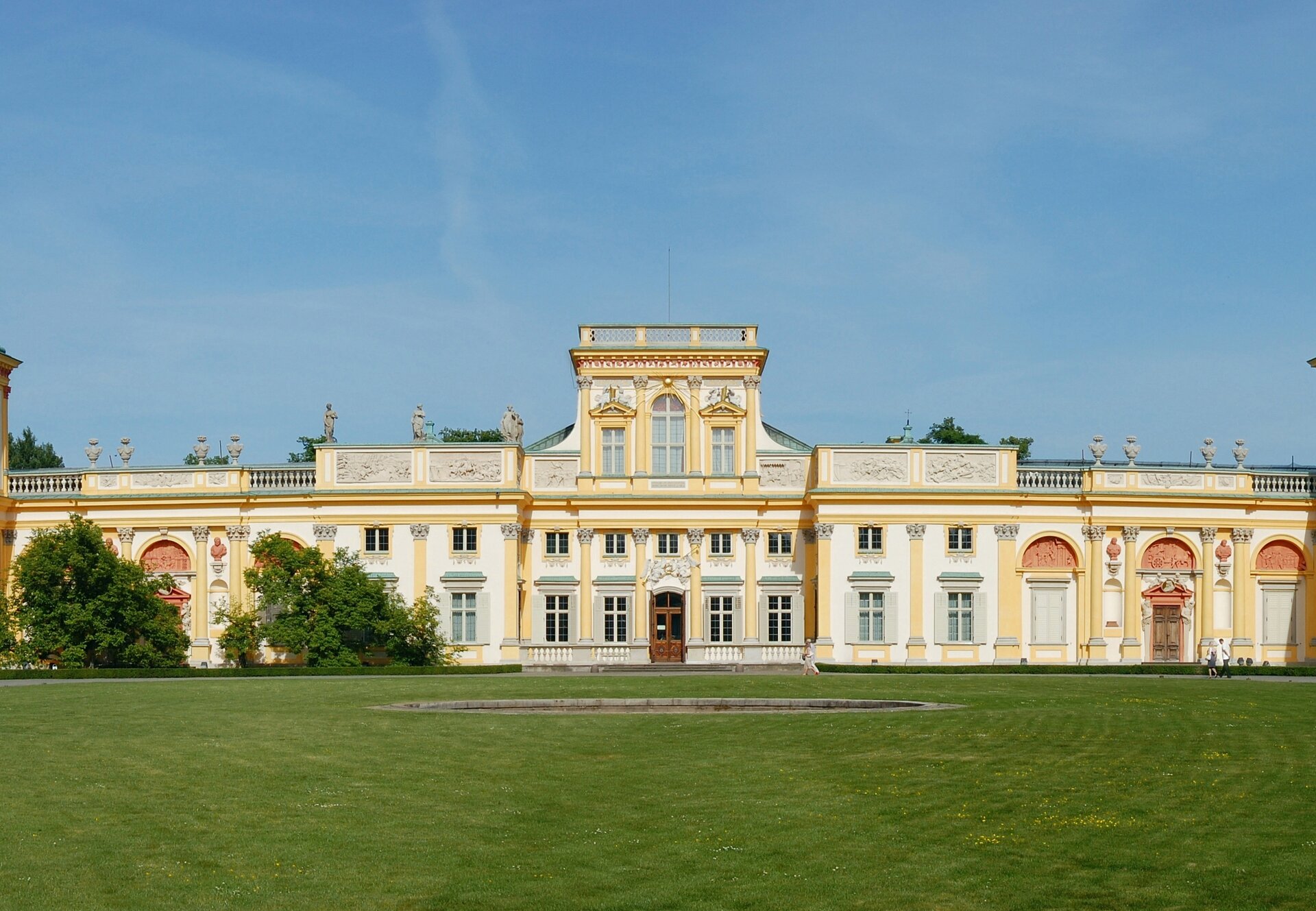 Ilustracja przedstawia fragment pałacu w Wilanowie. na obrazie widzimy fasadę budynku z wejściem głównym. Fasada jest koloru biało-żółtego. Przed budynkiem widoczna jest zielona trawa.