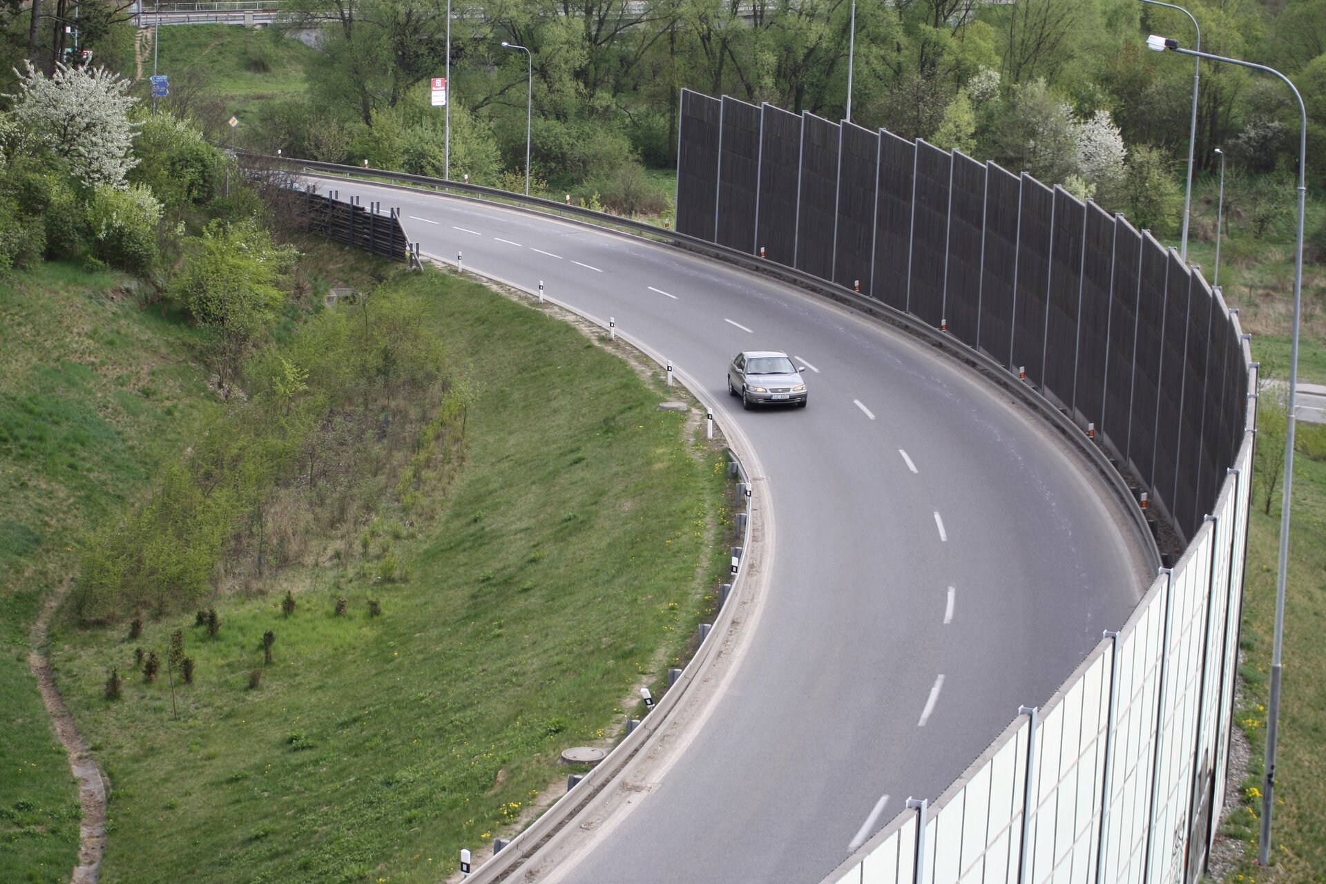 Zdjęcie przedstawia widok z lotu ptaka na dwupasmową drogę. Na drodze znajduje się samochód. Po prawej stronie drogi jest ekran akustyczny. 