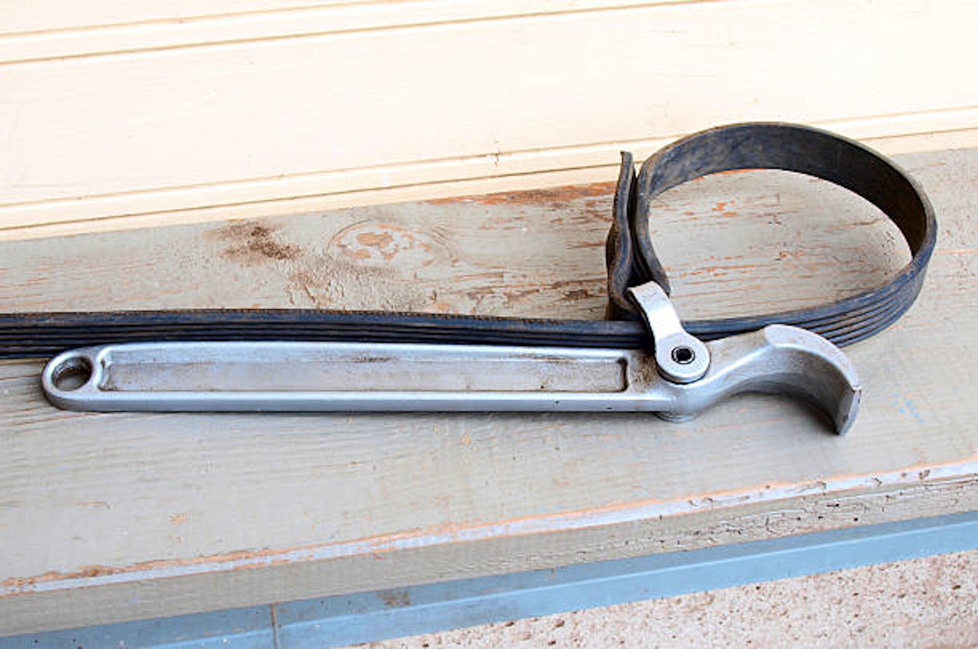 Na zdjęciu widoczny jest klucz taśmowy, długie i płaskie narzędzie wykonane ze stali. Jego część górna jest lekko zakrzywiona. Na klucz przymocowany jest pas gumowy za pomocą śruby, który tworzy pętlę.