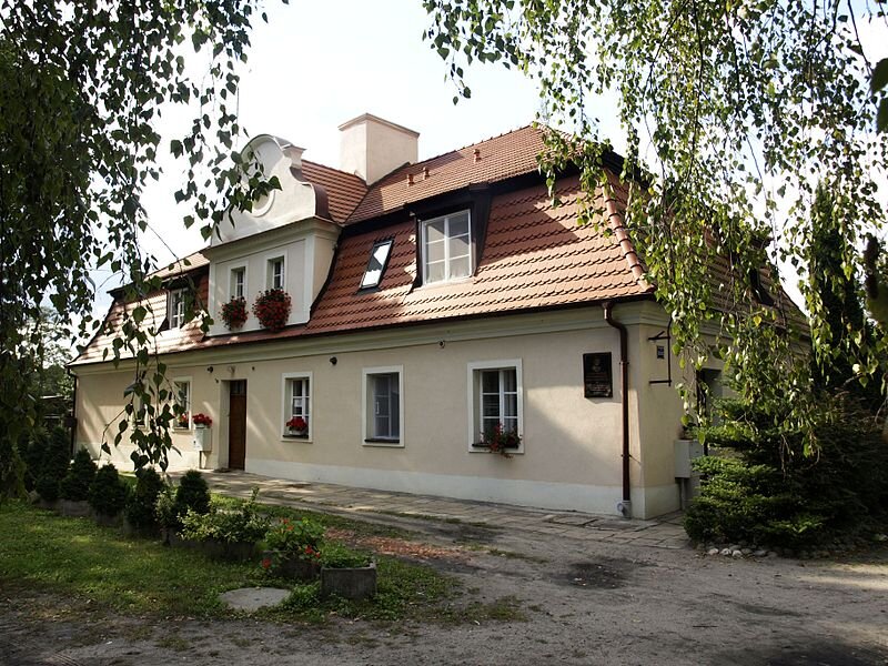 Zdjęcie przedstawia parterowy budynek z przodu z prawą boczną ścianą. Dom ma kształt prostokąta z poddaszem, w którym umieszczona jest lukarna, gdzie znajduje się okrągłe okno w górnej części. W dolnej części są dwa prostokątne okna ozdobione kwiatami. Dach pokrywa czerwona dachówka, gdzie w centrum znajduje się wysoki, prostokątny, biały komin. Elewacja ma jasny kolor. Na parterze znajdują się prostokątne, duże okna, ułożone obok siebie. Na zewnętrznych parapetach stoją czerwone kwiaty. Pośrodku budynku jest umiejscowione wejście. Przy prawej krawędzi frontowej ściany wisi czarna prostokątna tablica. Obok domu rosną świerki. Przed budynkiem znajdują się kwiaty i młode tuje w donicach.