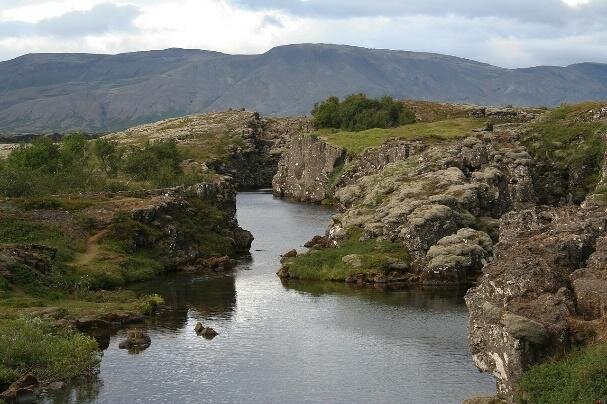 Na fotografii podłużna szczelina o szerokości kilkunastu metrów między pionowymi skalnymi ścianami, które pokryte są głównie porostami i trawą. W tle masywne wzniesienie.