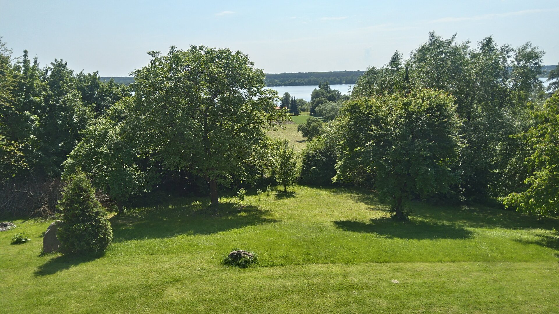 Fotografia przedstawia łąkę, na niej po prawej i lewej stronie kępy drzew. Na pierwszym planie kamienie ułożone jeden na drugim. Za nimi płot. W tle rozległa rzeka. 