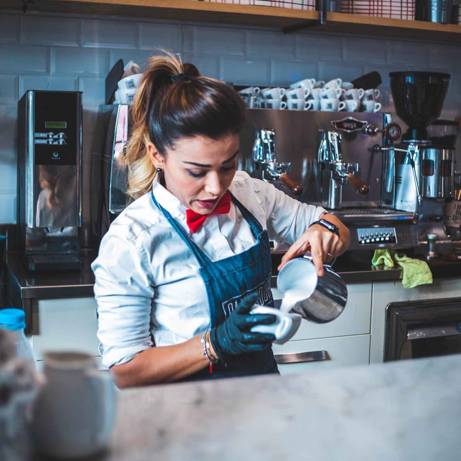 Zdjęcie przedstawia kobietę za ladą. Kobieta jest ubrana w białą koszulę, czerwoną muszkę i fartuch ochronny. W lewej ręce trzyma przechylony dzbanek z mlekiem, które wlewa do małej filiżanki. W tle jest ekspres do kawy, stos filiżanek ułożonych jedna na drugiej i wyciskarka do soków.