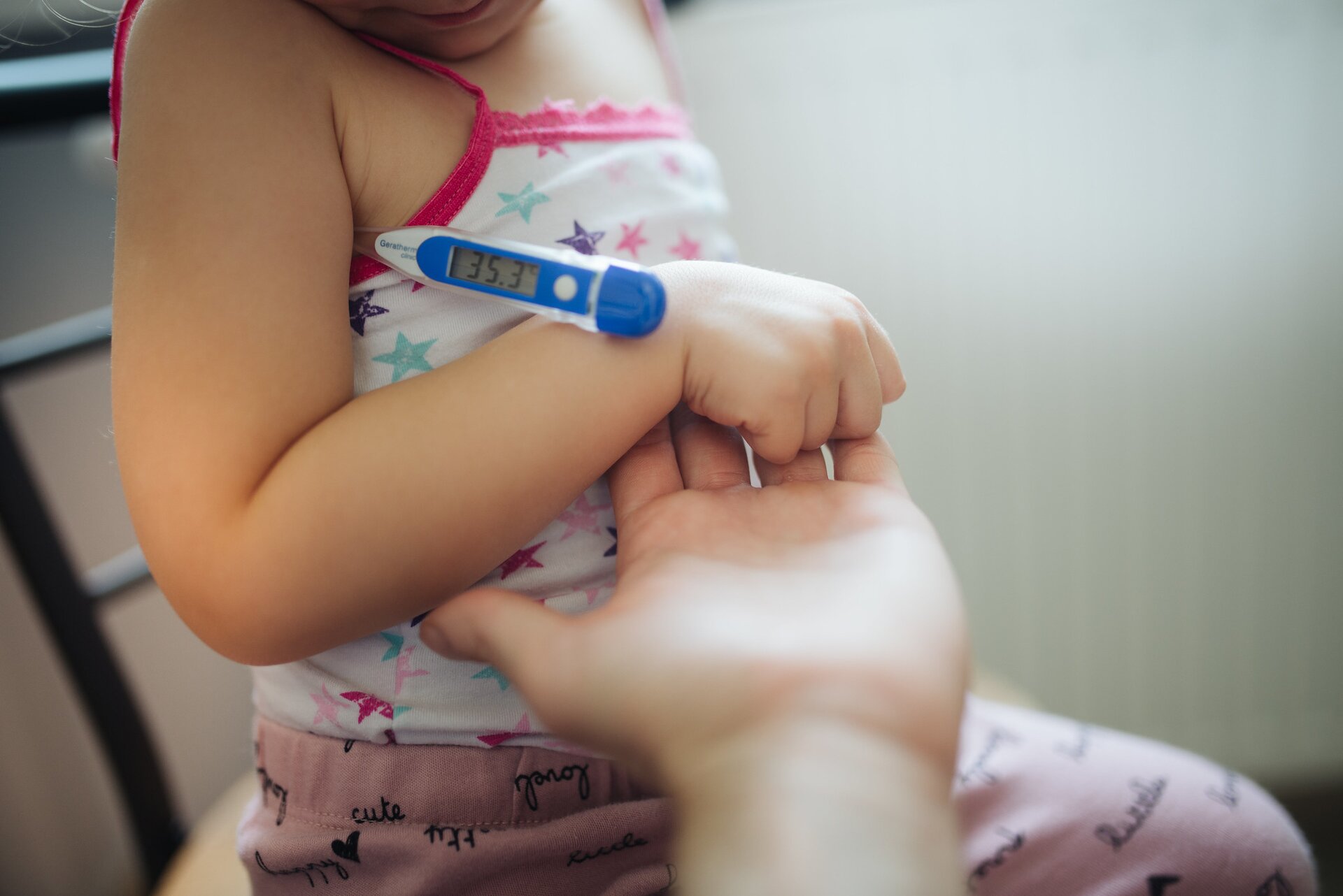 Ilustracja przedstawia dziewczynkę siedzącą na krześle. Widać jej tułów i jedno ramię. Pod pachą trzyma termometr elektroniczny. Węższa końcówka schowana jest pod pachą, widać tylko szerszą część urządzenia z guzikiem i ekranem elektronicznym.