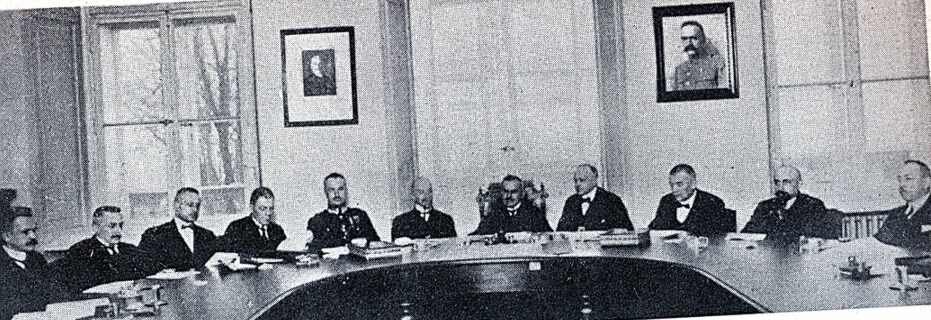 Na fotografii jest fragment owalnego stołu, przy którym siedzi jedenastu mężczyzn. W centrum nich jest Władysław Grabski. Za mężczyznami są okna i ściany, na których wiszą dwa portrety. Jeden z nich przedstawia Józefa Piłsudskiego. Na stole, przy każdym mężczyźnie są kartki.