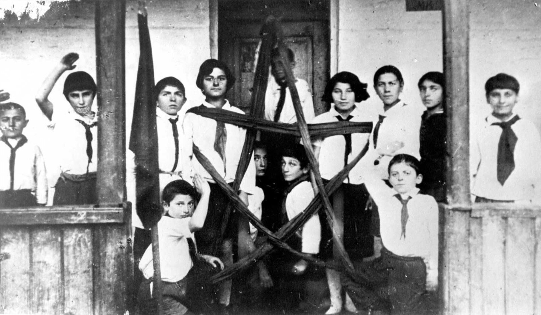 Fotografia czarno-biała przedstawiająca członków organizacji pionierskiej. Na ilustracji kilkoro dzieci stoi w sieni domu. Dziewczęta i chłopcy w wieku szkolnym. Wszyscy ubrani są w białe koszule, krawaty oraz czarne spodnie lub spódnice. Trzymają w dłoniach ułożony z taśmy znak pięcioramiennej gwiazdy.