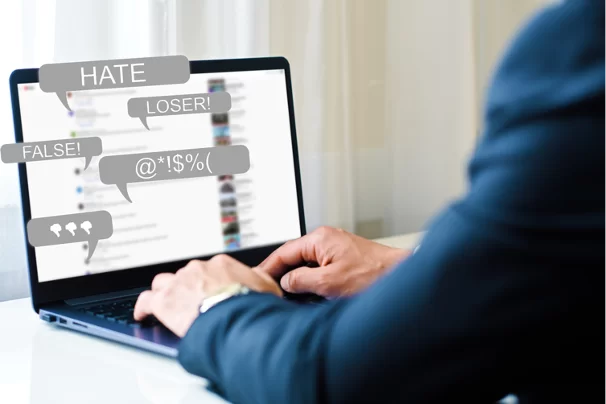 Ręce mężczyzny na klawiaturze laptopa. Pełny obraz na monitorze zamazany, w chmurkach dialogowych sugerujących treści pisane przez mężczyznę napisy po angielsku: Hate, Loser!, False! oraz nieprzyjazne emotikony.