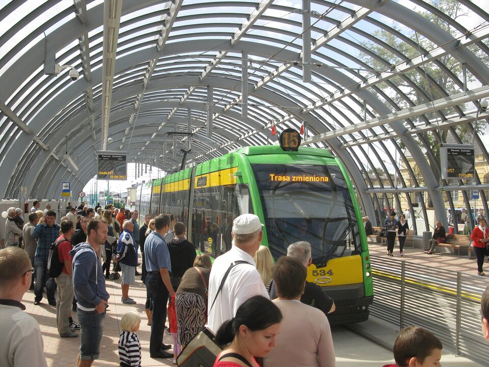 Zdjęcie przedstawia przystanek tramwajowy. Na przystanku stoi dużo ludzi. Nad przystankiem jest półokrągła wiata zrobiona z metalowej konstrukcji.   