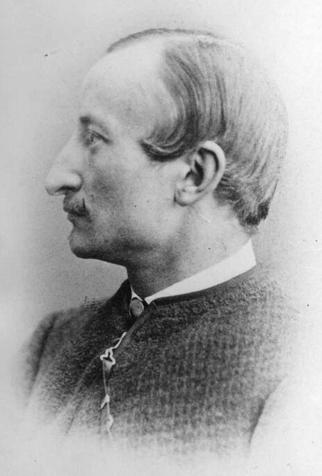 Ilustracja przedstawia portret mężczyzny w sile wieku. Postać ukazuje lewy profil. Mężczyzna ma krótkie, proste włosy zaczesane na prawą stronę. Mężczyzna ma wysokie, gładkie czoło. Jego brwi i oczy są wyraziste. Patrzy przed siebie. Jego nos jest haczykowaty. Mężczyzna ma niewielkie wąsy i wąskie usta. Jego policzki i podbródek są gładko ogolone. Jest ubrany w koszulę i surdut.