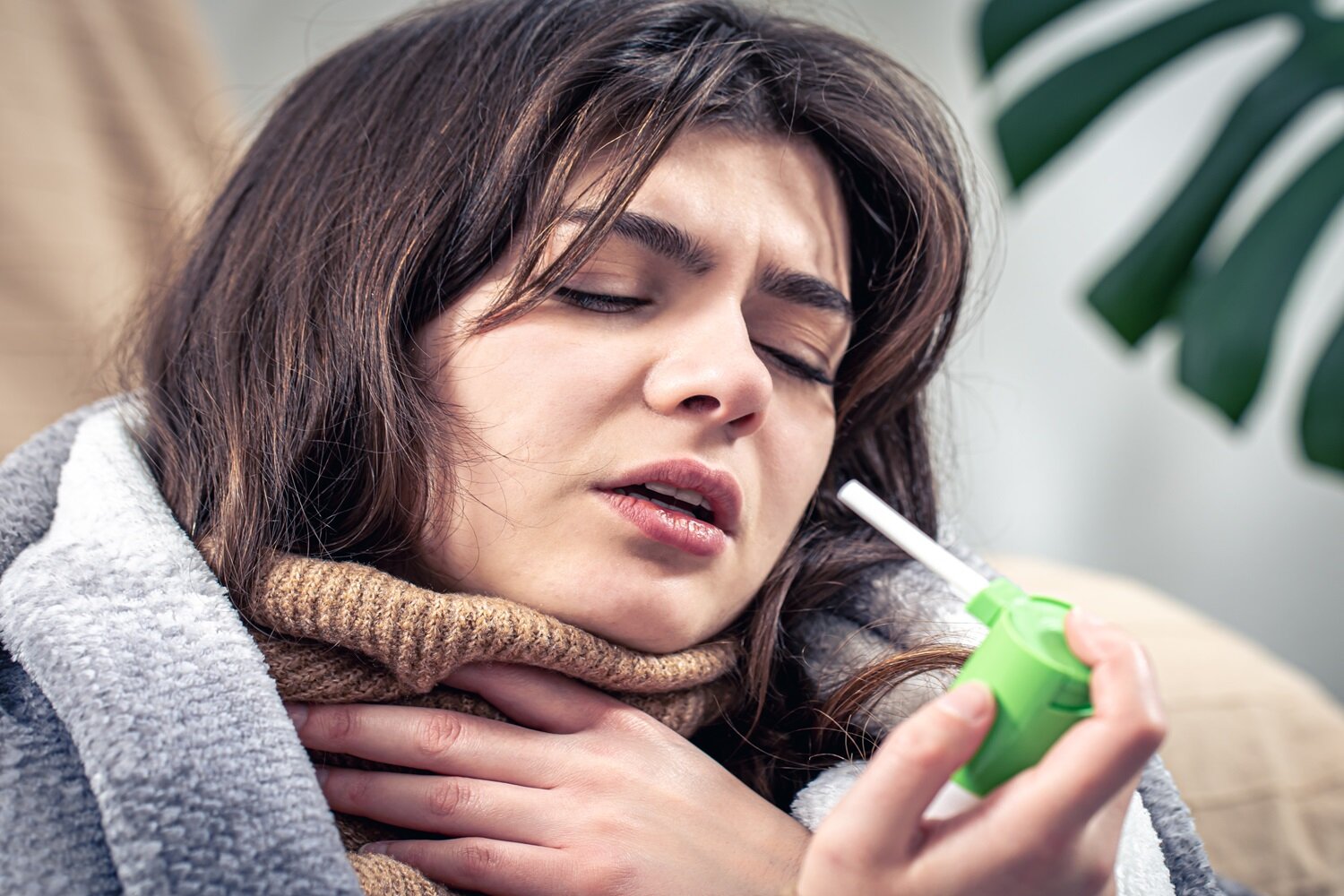 Zdjęcie przedstawia kobietę od ramion w górę, która lewą dłonią trzyma się za gardło, a w prawej trzyma małą butelkę z przytwierdzoną poziomo rurką. Kobieta ma ściągnięte brwi, przymknięte oczy, rozchylone usta, a jej głowa jest lekko przechylona do lewego ramienia.