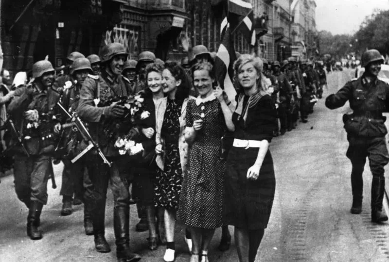 Kliknij, aby powiększyć Na zdjęciu niemieccy żołnierze maszerują ulicą. Są uśmiechnięci. Kilka młodych uśmiechniętych kobiet wita ich kwiatami. W tle widać kamienice i ludzi stojących na chodniku.