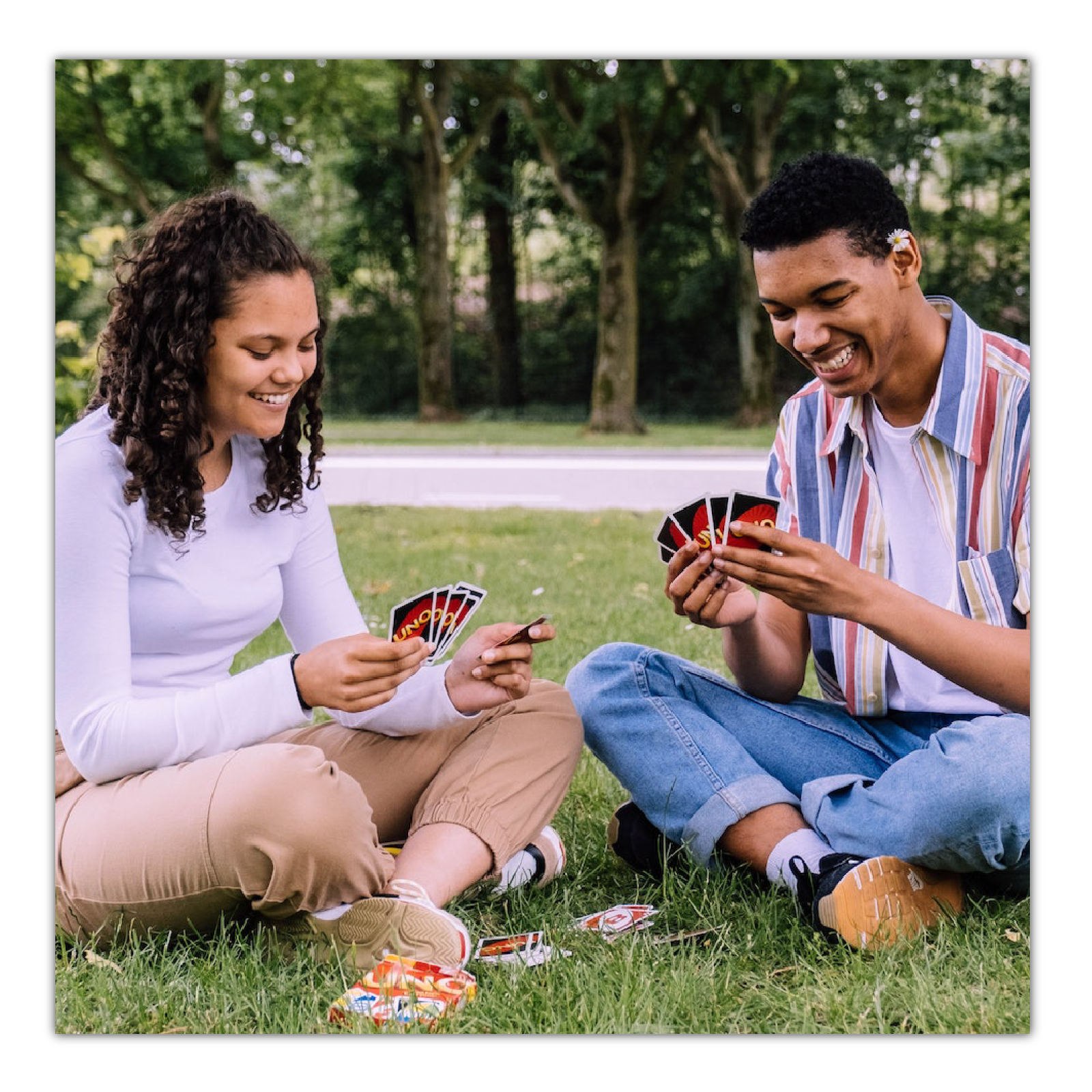 Ilustracja przedstawia chłopaka i dziewczynę siedzących na trawie i grających w karty uno.