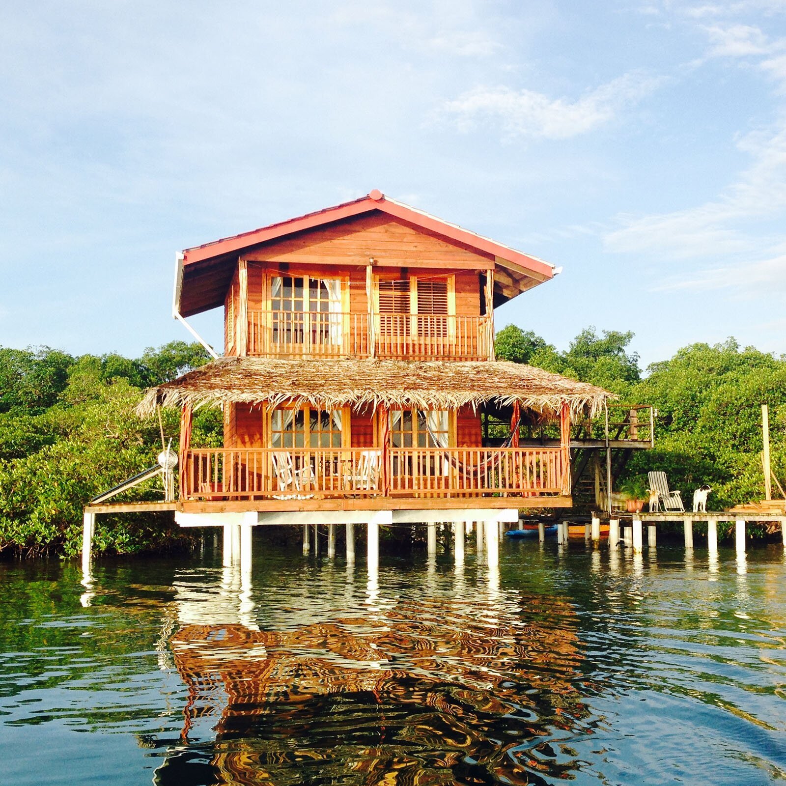 Zdjęcie przedstawia drewniany domek na wodzie, wybudowany na białym, szerokim molo. Wznosi się on nad akwenem wodnym, za którym widać gęstą, soczysto zieloną dżunglę. Domek jest piętrowy. Na parterze znajduje się dodatkowo taras, zadaszony przy wykorzystaniu wysuszonych liści egzotycznych roślin. Na piętrze znajduje się mały balkon. Domek posiada dwuspadowy, czerwony dach. 