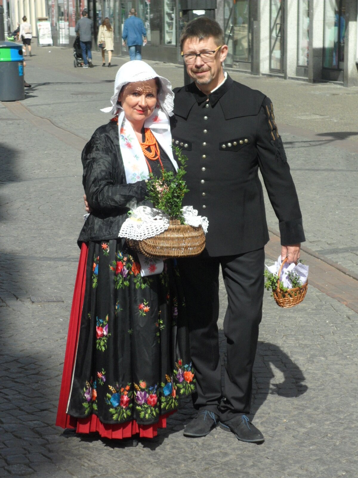 Zdjęcie przedstawia kobietę i mężczyznę w tradycyjnych strojach, w dłoniach trzymają plecione koszyczki z wielkanocną święconką. Kobieta ubrana w długą, czarną suknię z motywami kwiatowymi, na szyi korale, nosi czarną kurtkę, na głowie biały kapelusz. Mężczyzna z w czarnym mundurze górniczym. Długie proste spodnie, marynarka ze srebrnymi guzikami.