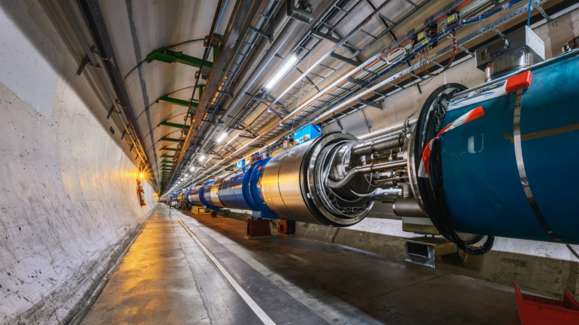 Rys. 6. Na zdjęciu widoczny jest fragment tunelu Wielkiego Zderza Hadronów. Tunel w przekroju jest kołem. W prawej części tunelu znajduje się metalowa rura, w której przyspieszane są cząstki. Na rurze znajduje się bardzo dużo przewodów biegnących wzdłuż niej. W tunelu widoczne są instalacje elektryczne i sieć kabli ciągnąca się wzdłuż sufitu. Jest to to samo zdjęcie, które było pokazane we wprowadzeniu.