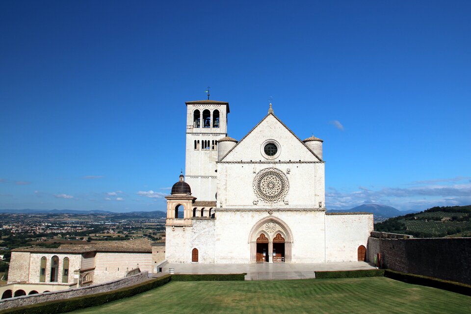 Ilustracja o kształcie poziomego prostokąta przedstawia fotografię Bazyliki Świętego Franciszka w Asyżu. Widok ukazuje bryłę kościoła od strony zachodniej. Dwukondygnacyjna bryła zakończona jest trójkątnym przyczółkiem. Podwójne wejście zwieńczone jest trójliściem i rozetą pomiędzy drzwiami. W przyczółku znajduje się okrągłe okno. Po lewej stronie widoczna jest niewielka wieża dzwonnicza. Dalej masywna, wyższa wieża z dwoma rzędami okien. Plac przed kościołem porośnięty jest trawą i otoczony murami. W dali widoczna jest panorama miasta. 