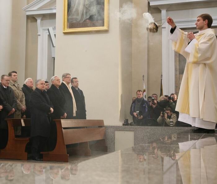 Na zdjęciu wnętrze kościoła. Z prawej strony ksiądz w złotej szacie sakralnej z kadzidłem. Przed nim ławki, a w nich stoją mężczyźni ubrani w czarne płaszcze. Pomiędzy ławami, a podwyższeniem, na którym stoi ksiądz, stoją mężczyźni trzymający aparaty.