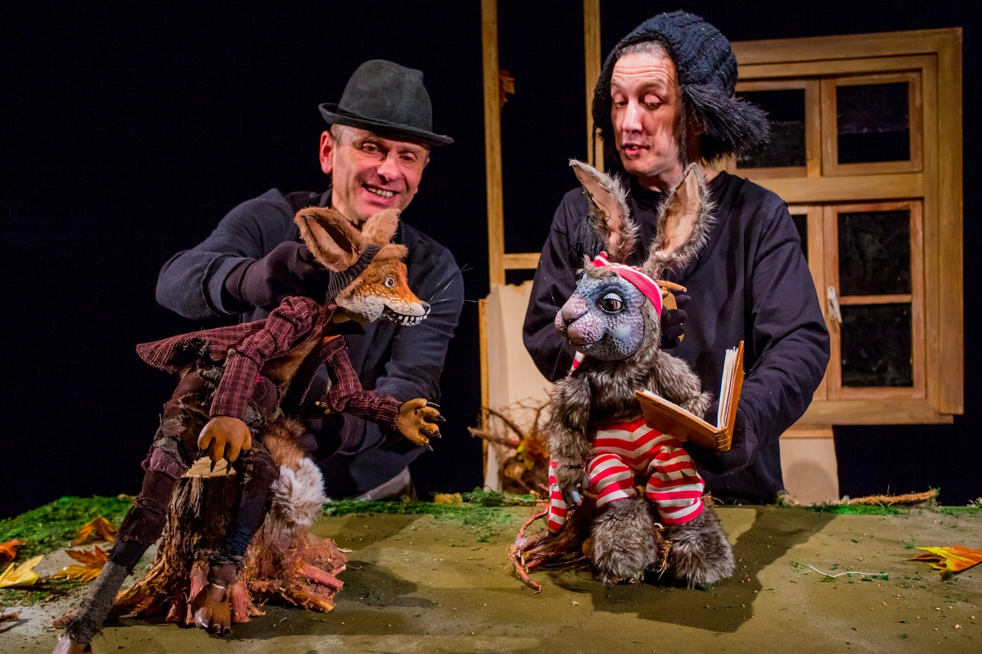 Ilustracja przedstawia fragment spektaklu Teatru Lalek „Banialuka” pt. „Smacznego, proszę wilka!”. Na scenie widać dwóch aktorów w czarnych kostiumach. Jeden nosi kapelusz, drugi czapkę uszankę, obaj animują lalki teatralne lisa i zające. Rudy lis ubrany jest w bordowy frak, ma ostre pazury. Zając trzyma w ręku książkę, nosi spodnie w biało‑czerwone poziome pasy. Tło jest czarne, na nim umieszczono drewniane okno.