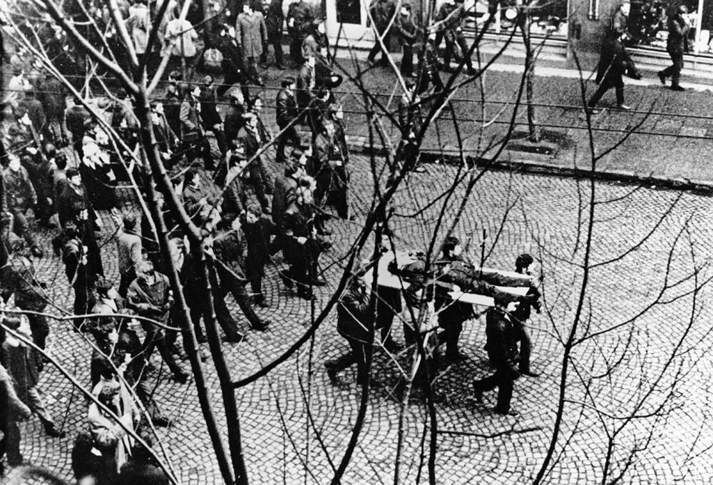 Czarno-biała fotografia przedstawiająca protesty w grudniu 1970 w Gdyni. Ulicą przechodzi tłum ludzi. Na czele demonstranci niosą na drzwiach ciało Zbyszka Godlewskiego.