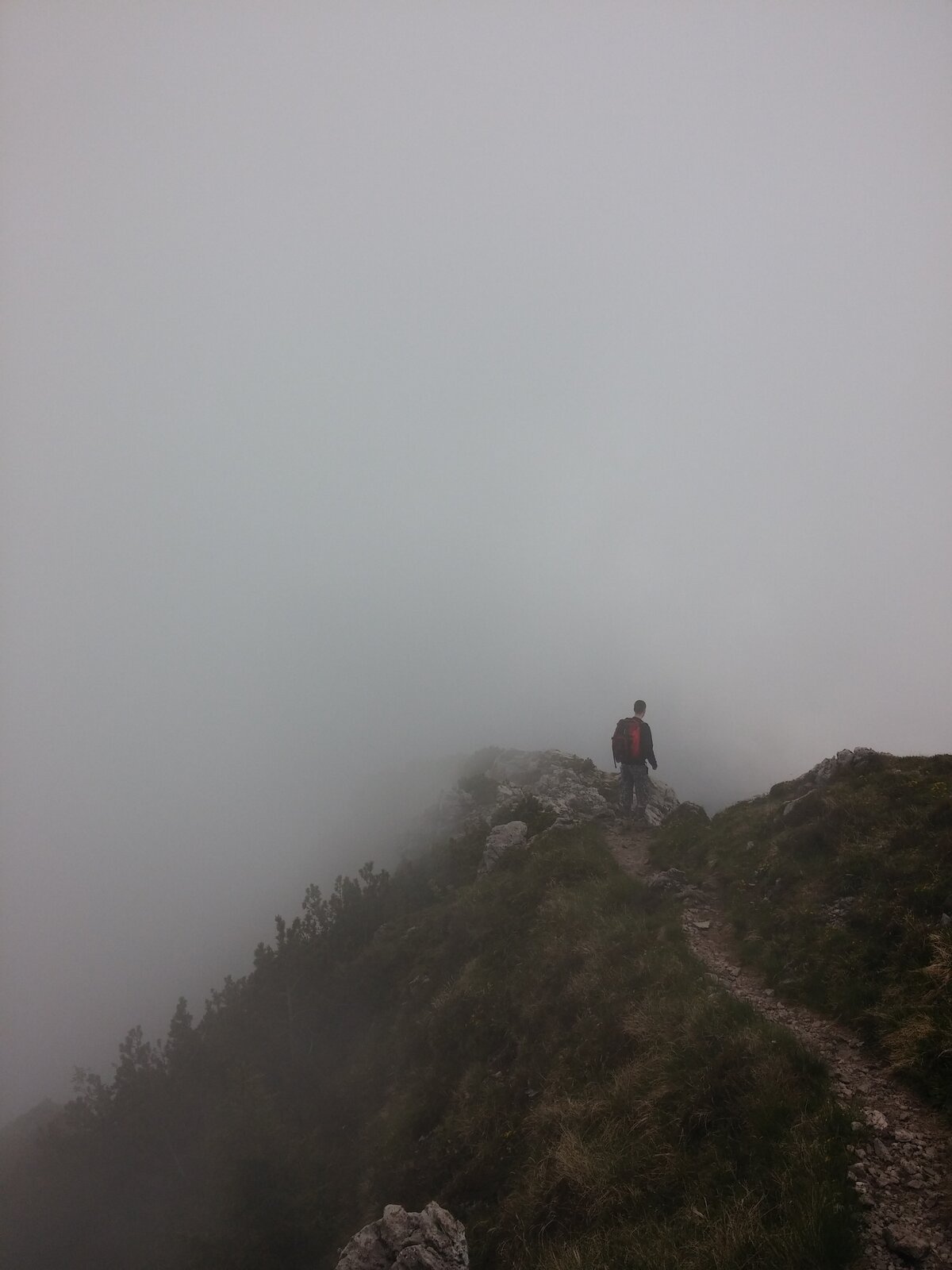 Na zdjęciu widać mężczyznę idącego szlakiem w górach, przed nim gęsta mgła. Turysta pieszy zmierza wąską ścieżką. Wzniesienie jest porośnięte roślinnością.