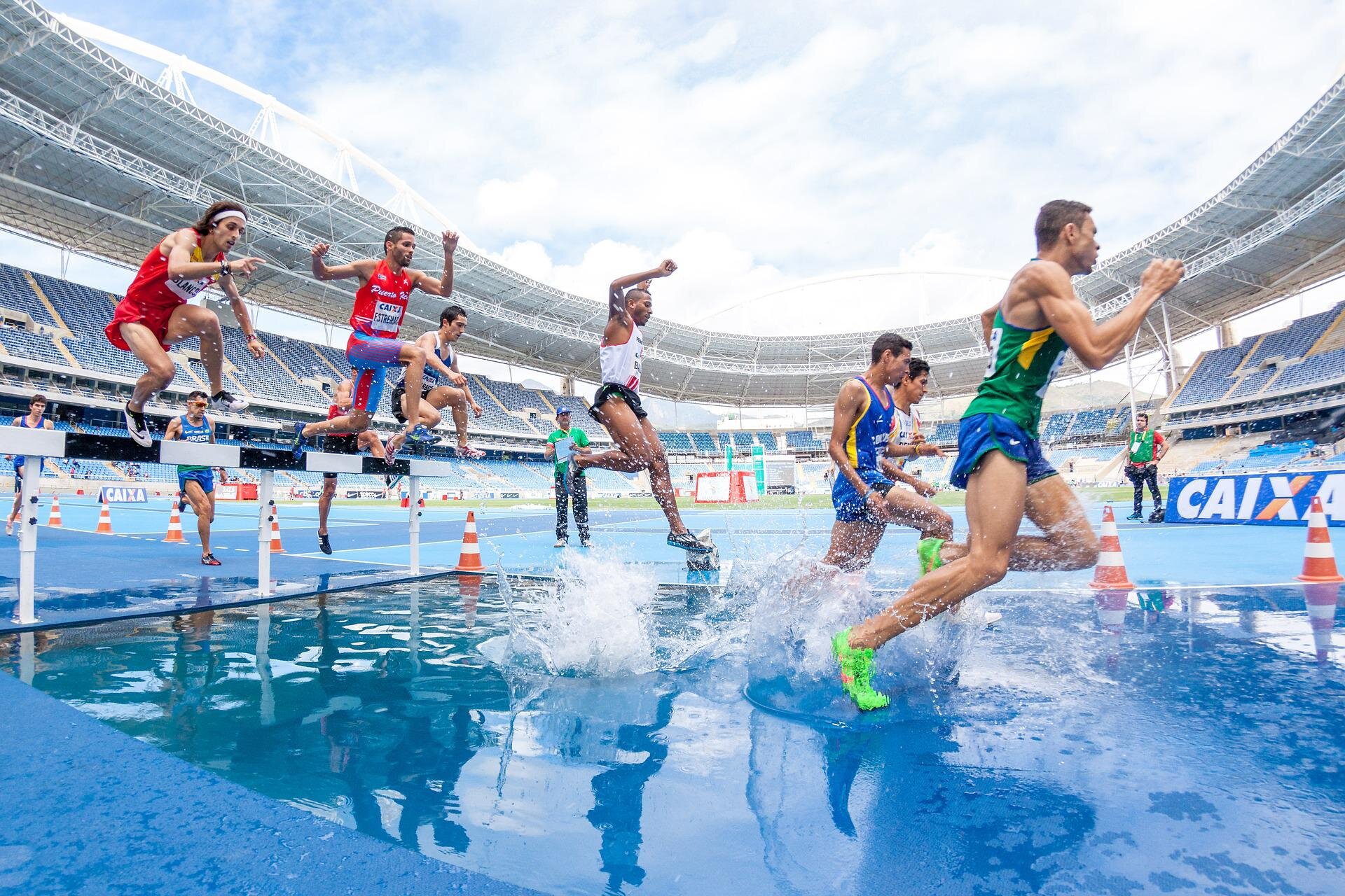 Zdjęcie przedstawia sportowców - biegaczy. Biegną na stadionie. Uchwycono moment, w którym część biegaczy jest nad płotkiem, a część biegnie w płytkiej wodzie. 