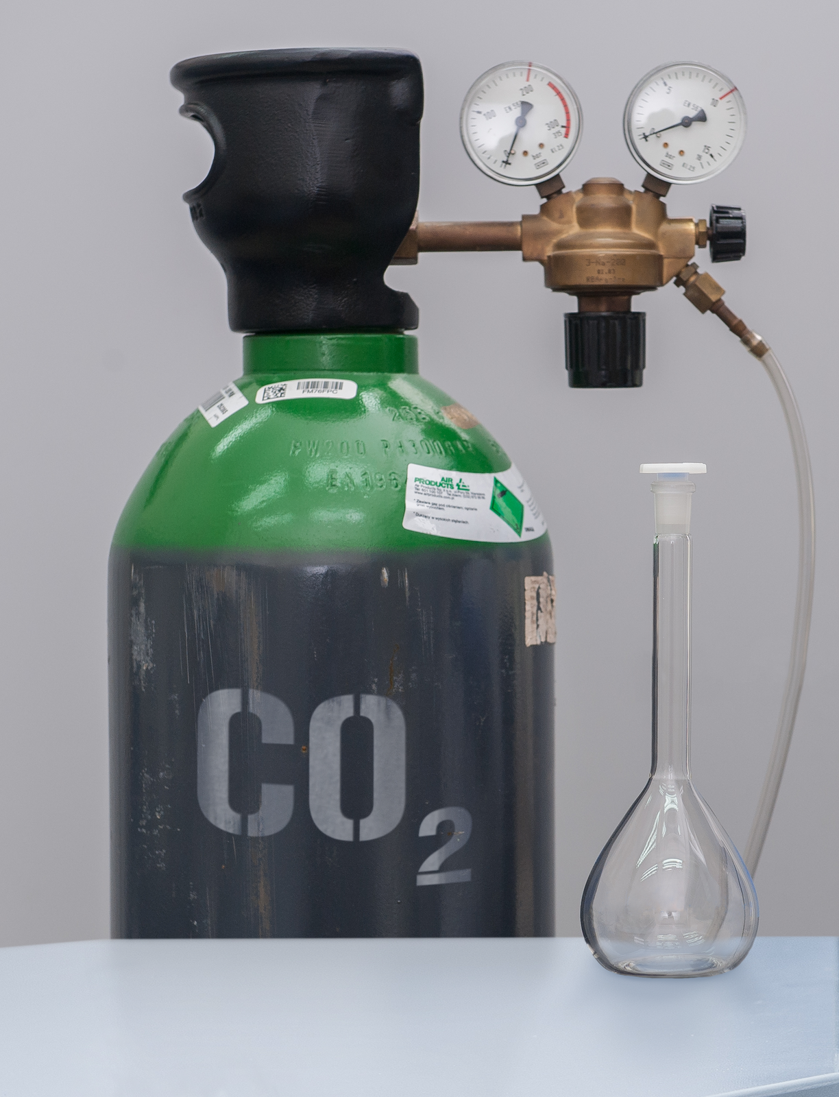 Zdjęcie przedstawia stolik z przezroczystym zamkniętym naczyniem szklanym. Za nim stoi butla z gazem zaopatrzona w zawór opisana symbolem dwutlenku węgla C O indeks dolny, dwa, koniec indeksu dolnego.