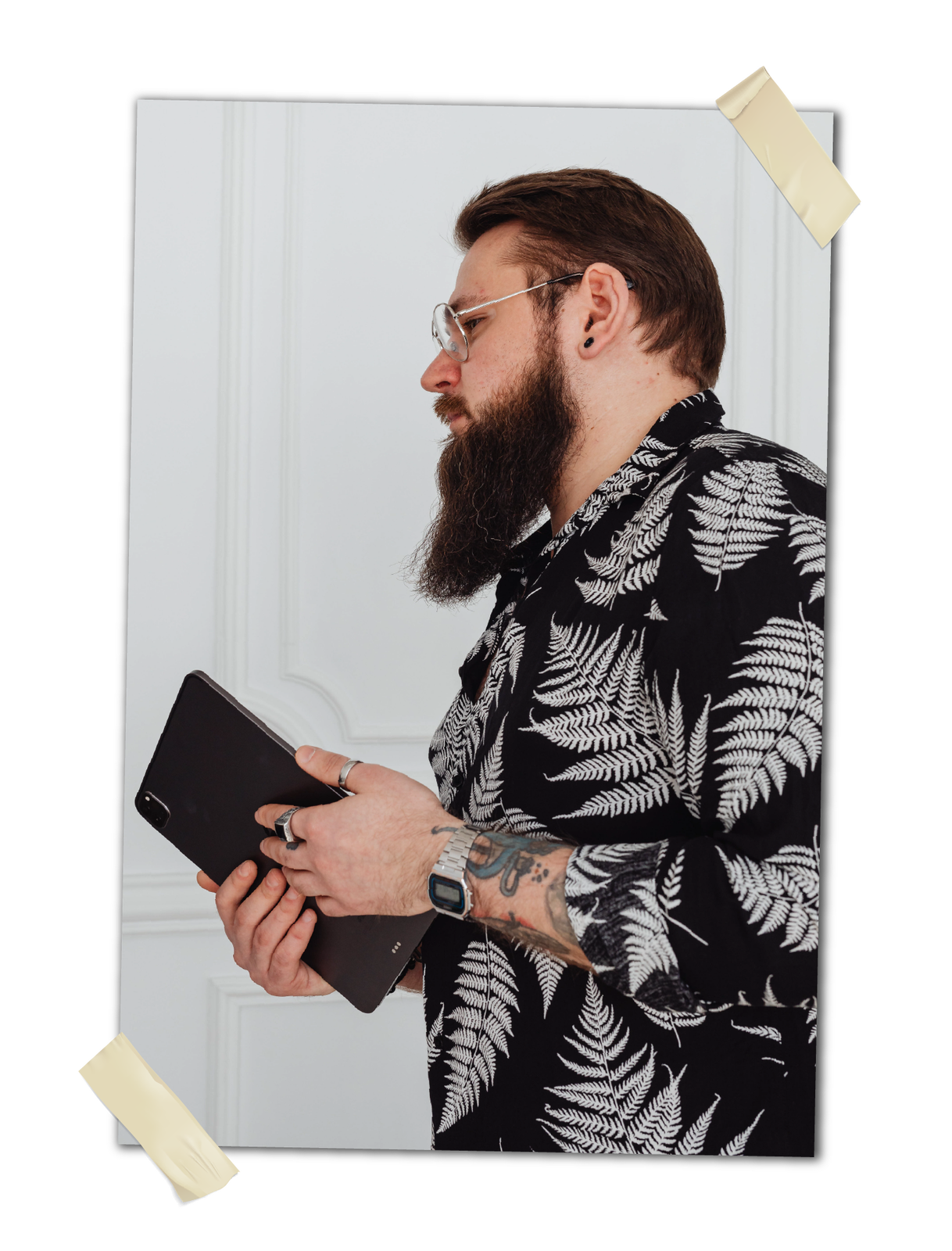 Zdjęcie przedstawia mężczyznę z długą, brązową brodą i wąsami, noszącego okulary. Jego brązowe włosy są przycięte i zaczesane do tyłu. W rękach trzyma tablet. Na nadgarstku widać zegarek, a spod koszuli wystaje tatuaż na jego ręce.