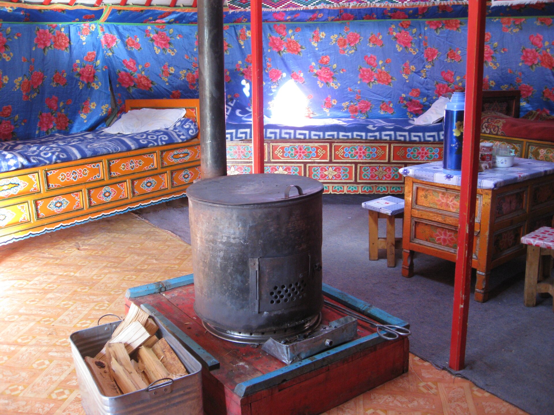 Zdjęcie przedstawia wnętrze jurty. Na środku palenisko z ogromnym garnkiem. Z tyłu drewniane łóżko, stolik i dwa krzesła.