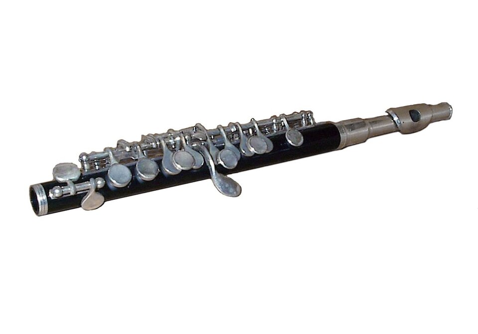 Flet piccolo. Zdjęcie przedstawia flet piccolo. Instrument ma krótki, wąski, czarny korpus i srebrny mechanizm z licznymi klapami. Leży ukośnie na białym tle. Po prawej stronie widać srebrną główkę z otworem zadęciowym, a po lewej zakończenie instrumentu.