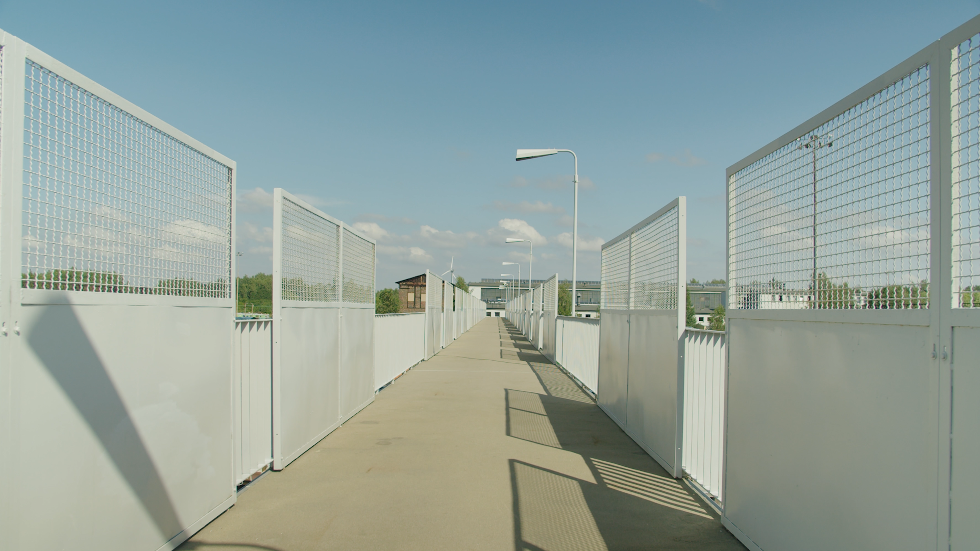  Ilustracja przedstawia przejście na kładce dla pieszych, która znajduje się ponad torowiskiem. Przejście wykonane jest z betonu, a obok znajdują się barierki zabezpieczające. Co jakiś czas na barierkach zamontowane są płoty w kształcie prostokątów, z częścią wystającą poza barierkę zbudowaną z metalowej siatki. W tle widoczne są latarnie oświetlające przejście, a na dalszym planie widać niebo oraz chmury. Kładka umożliwia pieszym bezpieczne przekroczenie torów kolejowych.