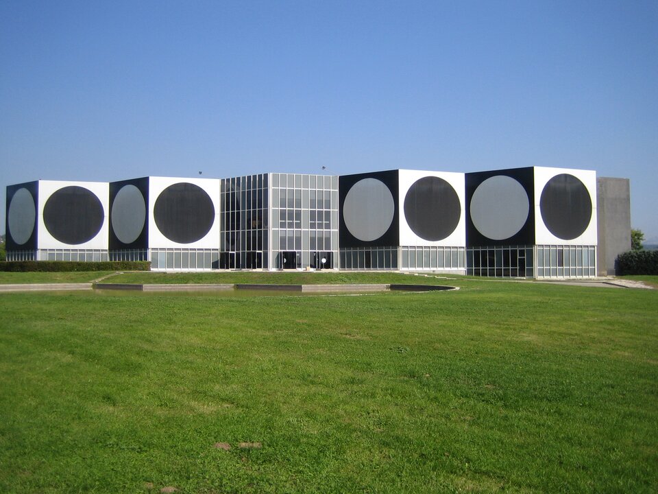 Ilustracja przedstawia muzeum w Aix‑en‑Provence we Francji, poświęcone twórczości Wiktora Vasarely'ego. Budynek kształtem przypomina składaną harmonijkę. Na kwadratowych ścianach muzeum widoczne są wielkie koła, białe na czarnym tle lub czarne na białym tle.