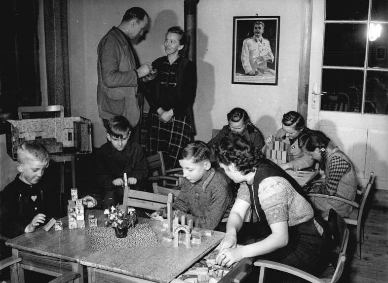 Zdjęcie przedstawia pomieszczenie. Na pierwszym planie przy stole siedzą dzieci, które bawią się klockami. Za nimi stoi drugi stół, przy którym druga grupa dzieci robi to sami. Po lewej stronie stoi mężczyzna i kobieta. Rozmawiają ze sobą. Na ścianie wisi portret Józefa Stalina w jasnym mundurze.