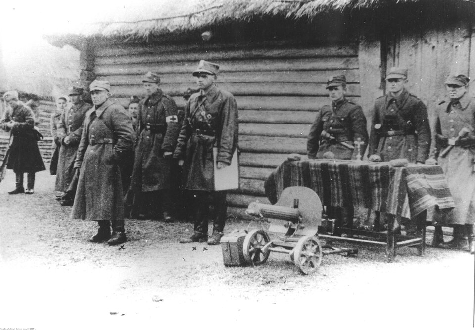 Czarno‑białe zdjęcie przedstawia grupę mężczyzn. Ubrani są w wojskowe płaszcze. Na głowach mają wojskowe czapki. Wszyscy stoją. Jeden z nich, stojący na środku fotografii, ma przewieszony przez ramię mapnik, czyli pokrowiec na mapę. Przed trzema mężczyznami z prawej strony stoi stół z obrusem, a przed stołem jest małe działo armatnie. Na stole stoi krzyż. W tle jest ściana drewnianego budynku.