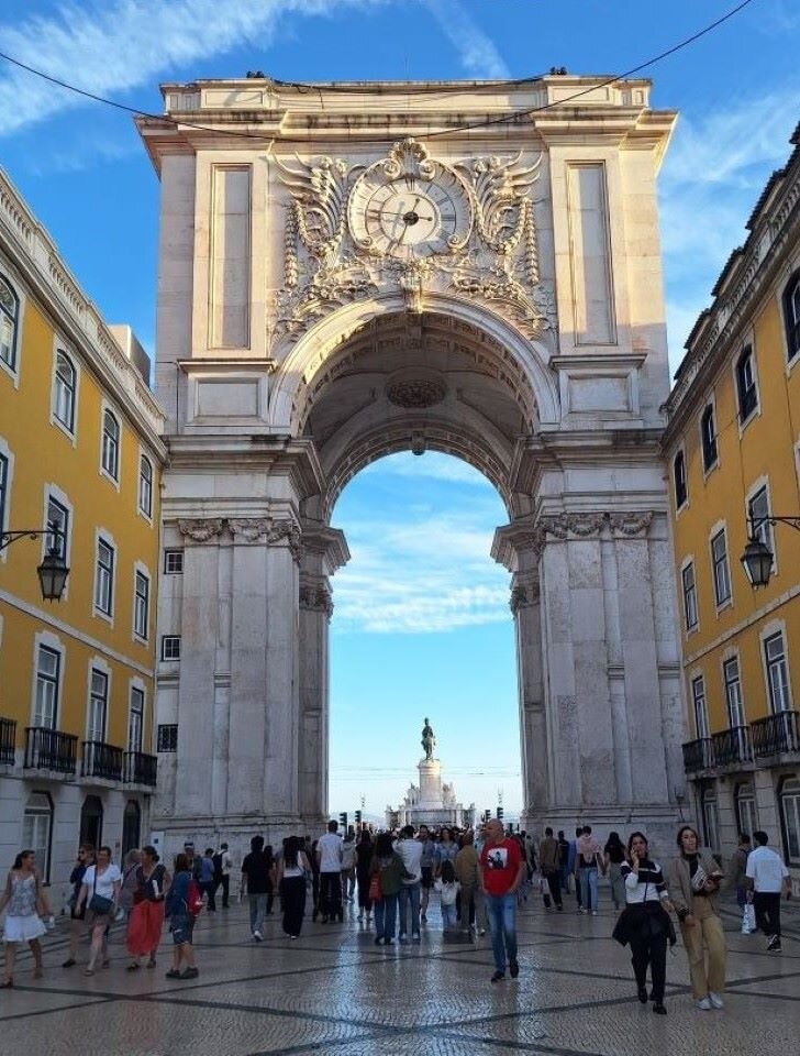 Ilustracja przedstawia fotografię miasta. W kadrze znajduje się fragment deptaku, po którym spacerują ludzie. Architektura miejska jest ujęta w kompozycji symetrycznej: pośrodku monumentalna brama, a po obydwu stronach przyległe do niej kamienice. W oddali, za bramą widoczny jest pomnik oraz błękitne niebo. Kompozycja kadru jest kompozycją otwartą.