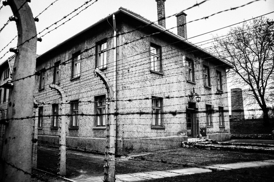 Zdjęcie przedstawia piętrowy, zbudowany z cegły budynek. Jest on otoczony drutem kolczastym.
