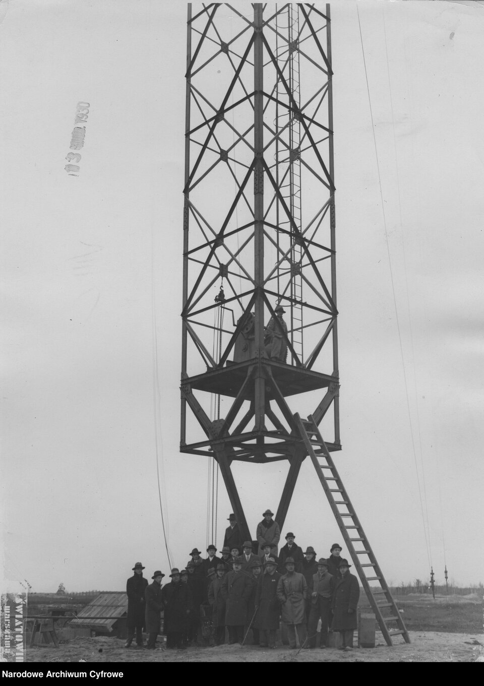 Czarno‑biała fotografia przedstawia grupę ludzi zgromadzonych u podstawy masztu radiowego w trakcie jego budowy lub inspekcji. Na pierwszym planie widoczna jest kilkudziesięcioosobowa grupa mężczyzn. Część osób znajduje się także na platformie konstrukcji. Centralnym punktem zdjęcia jest stalowy maszt o kratownicowej konstrukcji, sięgający wysoko poza górną krawędź kadru. Do platformy prowadzi drabina.